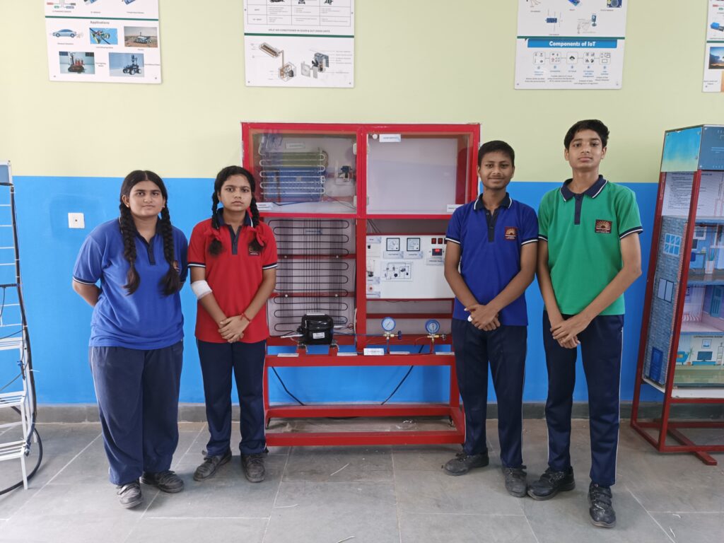 Four students in a vocational lab standing next to a Sagedel refrigeration and air conditioning trainer kit with visible compressor and gauges